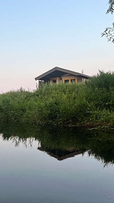 A quaint wooden cabin sits atop a grassy riverbank, reflected perfectly in the calm water below, surrounded by lush greenery under a clear, blue sky.