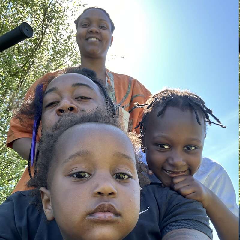 The image depicts a family of three posing for a selfie outdoors, with the father and two children visible, set against a backdrop of trees and a blue sky.