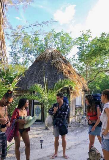 The image depicts a group of people standing outside a thatched-roof hut, surrounded by lush greenery and trees, with a serene blue sky above.
