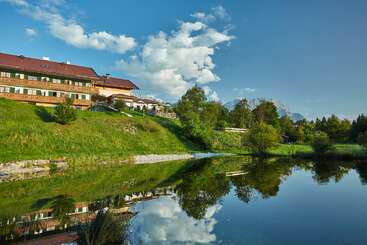 A large building with a red roof sits on a grassy hillside beside a calm lake, reflecting trees, blue sky, and distant mountains beautifully.