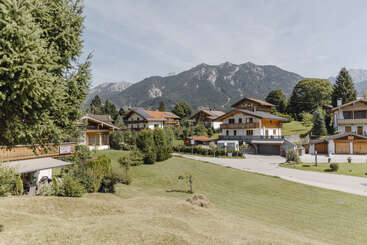 This image shows a picturesque mountain village with charming wooden houses, lush green lawns, trees, and a scenic mountainous backdrop under a bright, clear sky.