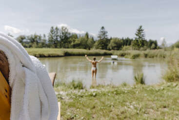 A person stands in a pond with arms raised, enjoying nature. In the foreground, a white towel is draped over a wooden surface near grass.