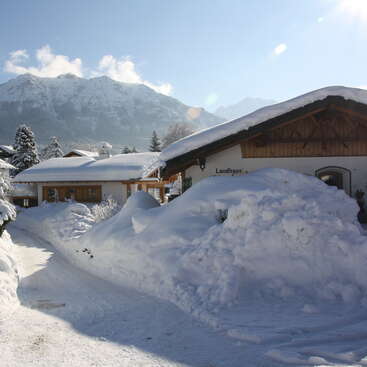 Charming alpine houses are covered in thick snow. Snowbanks line the path, mountains loom in the background, and bright sunlight glistens on a clear winter day.