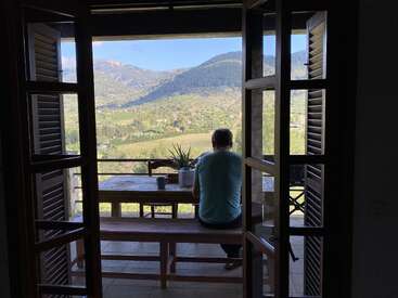 Uma pessoa senta sozinha em um banco na varanda, olhando para uma linda paisagem montanhosa através das portas abertas, curtindo a tranquilidade e a beleza natural lá fora.
