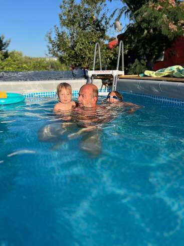A man swims in a blue above-ground pool with two young children, one sitting on his back. Surrounding are trees, blue sky, and pool toys.