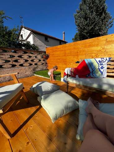 A sunny backyard with wooden deck, outdoor cushions, and furniture. A child in a diaper plays on green grass. Someone relaxes, feet up, enjoying the scene.