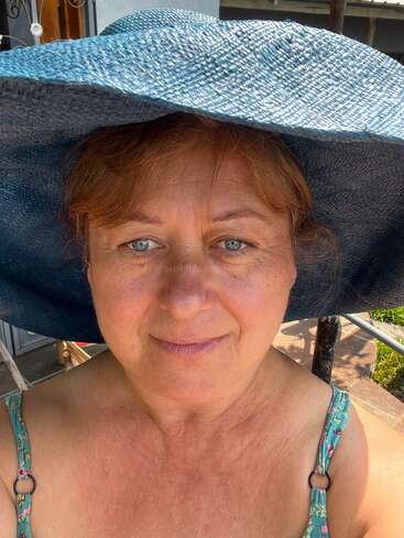 Das Bild zeigt eine Frau, die einen großen blauen Sonnenhut trägt, mit einem unscharfen Hintergrund aus einer Terrasse und einem Gebäude.