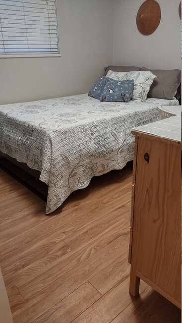 This is a cozy bedroom with wood flooring, a neatly made bed, decorative pillows, a small dresser, and simple wall decor. Light from a window.
