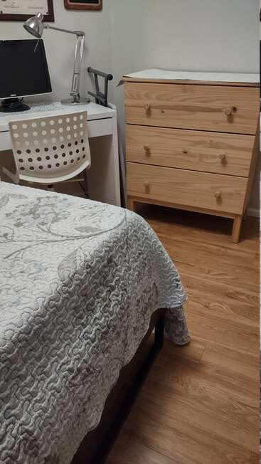 A cozy room with a neatly made bed, a white desk with a lamp, computer monitor, white chair, and a wooden dresser on a wooden floor.