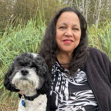 Une femme aux longs cheveux bruns sourit, assise à l'extérieur devant de hautes herbes, accompagnée d'un chien noir et blanc assis près d'elle.