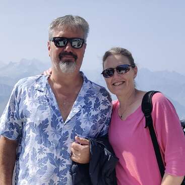 Um casal sorridente, ambos usando óculos escuros, posa junto ao ar livre. O homem veste uma camisa florida e segura um casaco, enquanto montanhas e o céu azul servem de pano de fundo.