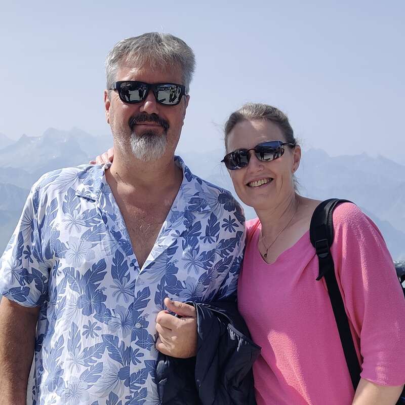 Um casal sorridente, ambos usando óculos escuros, posa junto ao ar livre. O homem veste uma camisa florida e segura um casaco, enquanto montanhas e o céu azul servem de pano de fundo.