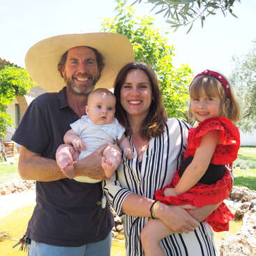 A happy family of four stands outside on a sunny day. The father wears a large straw hat and holds a baby, while the mother holds a smiling girl.