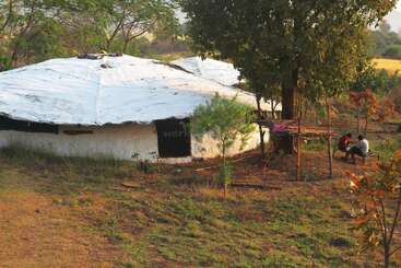The image depicts a serene rural scene with a white building, lush trees, and two individuals sitting on a bench, surrounded by a natural and peaceful environment.
