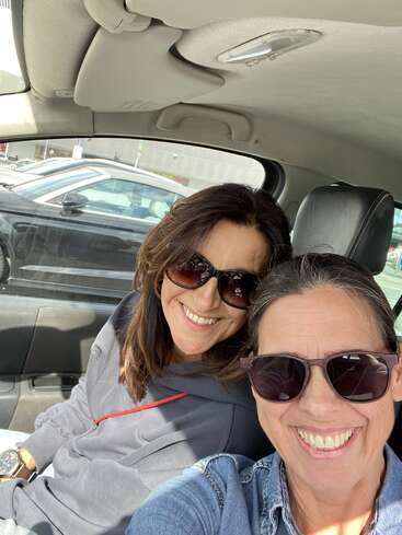 Two women are sitting in a car, both wearing sunglasses and smiling brightly. Sunlight streams in, highlighting their happiness on what appears to be a pleasant day.