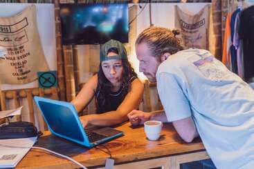 Two people, a woman in a backward cap and a man with a bun, are focused on a laptop at a wooden desk in a cozy, informal setting.