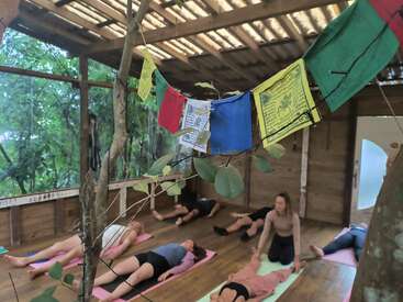 In a rustic wooden open-air room, people practice yoga on mats. Colorful prayer flags hang overhead, with green leafy branches adding a tranquil atmosphere.