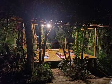 A peaceful outdoor yoga session at night, participants lying on mats under a wooden pavilion surrounded by lush tropical plants and soft, warm lighting.