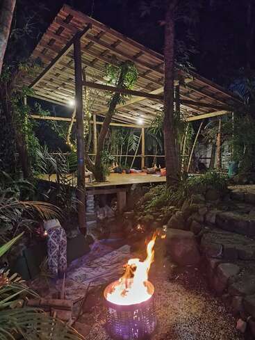 A cozy, open-air wooden pavilion in a lush jungle at night, softly illuminated. A fire burns in a metal drum, creating a warm, inviting atmosphere nearby.