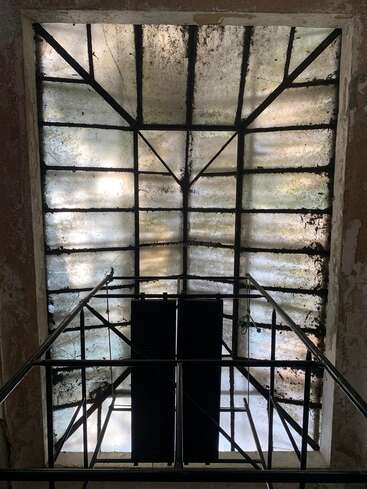 Un grand plafond de verre ancien et sale, avec un cadre métallique, partiellement recouvert de crasse. La lumière du soleil le traverse, illuminant le motif géométrique en damier et les ombres en dessous.