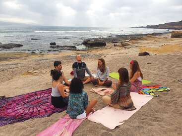 The image depicts a group of seven people sitting in a circle on the beach, with a serene ocean view and cloudy sky in the background.