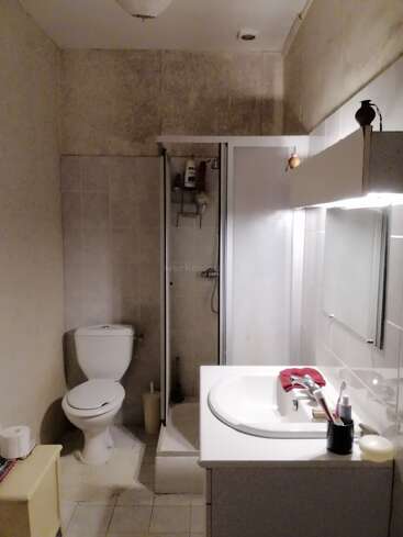 Esta é uma pequena casa de banho com vaso sanitário, box de chuveiro e pia. A bancada tem produtos de higiene pessoal e uma toalha vermelha; as paredes e o chão parecem simples e um pouco gastos.