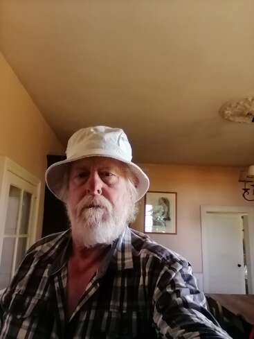 Um homem barbudo, usando um chapéu de pescador branco e uma camisa xadrez, está dentro de casa. O cômodo tem paredes em tom creme, quadros nas paredes, portas brancas e uma luminária no teto.