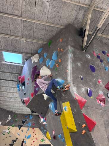 A person climbs an indoor bouldering wall, gripping colorful holds. The facility is spacious, with high ceilings and various climbing routes visible in the background.