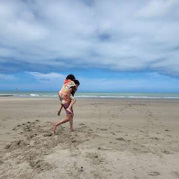 A person gives a piggyback ride to another on a sandy beach under a partly cloudy sky. The ocean is calm, with gentle waves and horizon visible.
