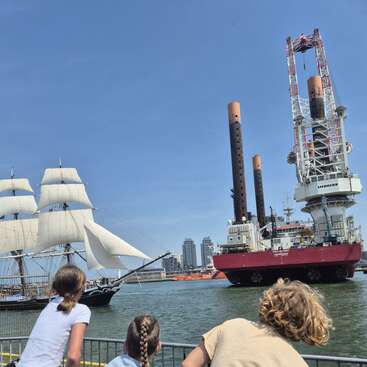 Derrière une clôture, trois enfants regardent un majestueux voilier et un grand navire industriel moderne se croiser sur un front de mer ensoleillé.