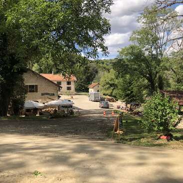 A peaceful rural scene with stone buildings, a cafe area with umbrellas, parked cars, greenery, trees, and a dirt path under a partly cloudy sky.
