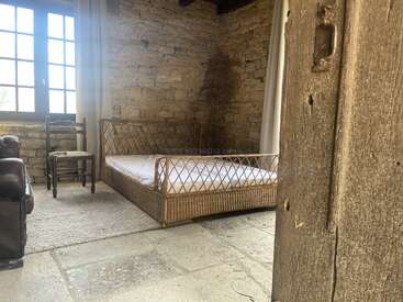 A rustic bedroom with stone walls, a wicker bed frame, large window, vintage chair, and cozy carpet. Natural light fills the serene, minimalist space.