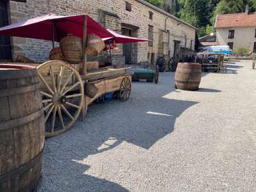 A rustic courtyard features old wooden barrels, carts with large wheels, red umbrellas, wicker baskets, and stone buildings under the bright sunny sky.