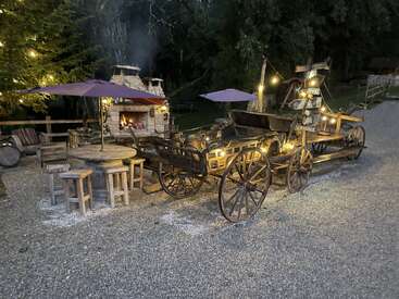 This cozy outdoor scene features rustic wooden carts with string lights, a stone oven fireplace, wooden stools, tables with umbrellas, and trees in the background, creating warmth.