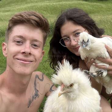 Dos personas sonrientes están sentadas sobre una exuberante pradera verde, cada una con una gallina Silkie blanca y esponjosa en brazos. Al fondo se ve un tranquilo paisaje rural con árboles y colinas.