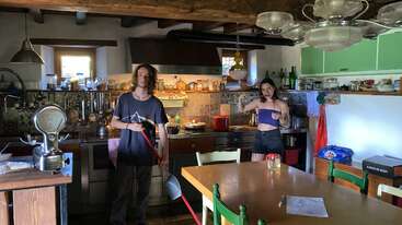Two young people in a cozy, rustic kitchen. One holds a mop, the other poses playfully. Sunlight shines in. Various kitchen items and food are visible.