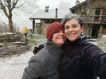 Two smiling women embrace for a selfie in a snowy yard, in front of a rustic house. Other people and trees are visible in the wintry background.