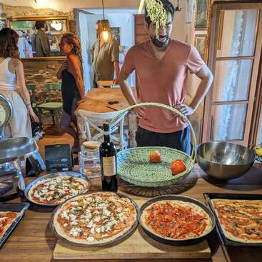 Das Bild zeigt eine geschäftige Küchenszene mit einem Mann, der hinter einem mit Pizzen beladenen Tisch steht, umgeben von Menschen, die sich im Hintergrund treffen.