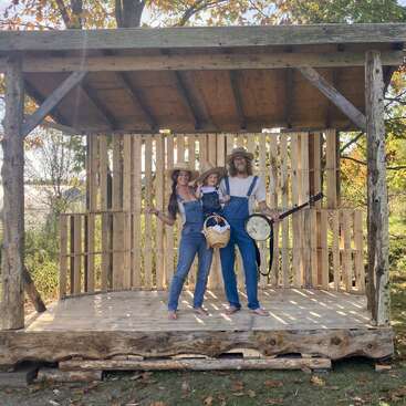 A family of three, dressed in overalls and straw hats, stands barefoot on a rustic wooden stage outdoors. One adult holds a banjo; it's autumn.