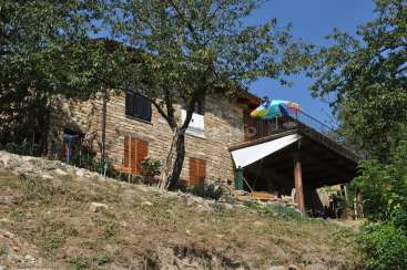 L'image représente une maison rustique en pierre avec un balcon, située sur une colline, avec un parapluie vibrant et entourée d'arbres luxuriants et d'un ciel bleu clair.