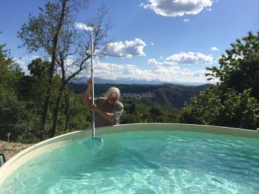 L'image représente un homme debout dans une piscine, tenant un skimmer, avec un paysage montagneux serein et des arbres en arrière-plan par une journée ensoleillée.