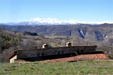L'image représente un bâtiment rustique en pierre avec un toit en tuiles, sur fond de collines et de montagnes ondulantes sous un ciel bleu clair.
