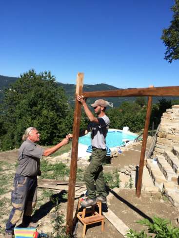 L'image représente deux hommes construisant une structure en bois dans une arrière-cour, avec une piscine et des arbres visibles à l'arrière-plan.