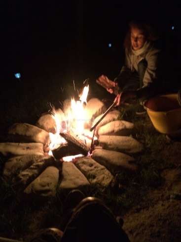 L'image représente une femme faisant rôtir un marshmallow sur un feu de camp, avec un seau à proximité, sur un fond sombre.