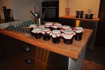 L'image représente un comptoir de cuisine avec des pots de confiture ou de conserves, un égouttoir à vaisselle et des ustensiles de cuisine, sur fond de cuisine avec un four et des casseroles.