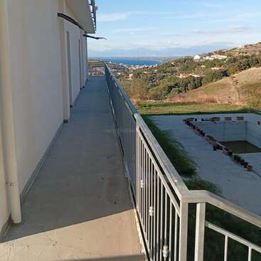 Ein langer Balkon mit einem Metallgeländer bietet einen tollen Blick auf eine malerische Landschaft mit Hügeln, Häusern und Wasser in der Ferne. Darunter ist ein leerer rechteckiger Bereich, vielleicht ein Pool.
