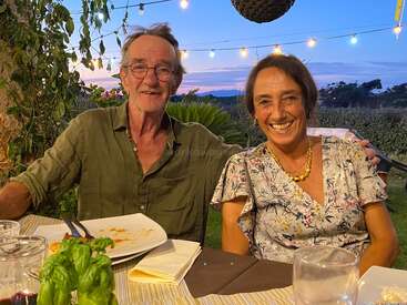 Um casal sorridente senta-se em uma mesa de jantar ao ar livre durante o pôr do sol. O ambiente é aconchegante, com luzes de corda, vegetação e pratos vazios, criando uma atmosfera calorosa e alegre.