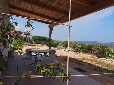 Um pátio coberto com uma mesa redonda, cadeiras brancas e plantas trepadeiras. Jardim com arbustos de flores coloridas, cachorro descansando e montanhas visíveis ao fundo. Cena tranquila.