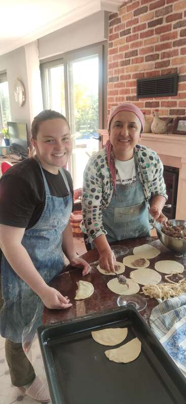 Two smiling women, wearing aprons, are making stuffed pastries together in a cozy kitchen. Sunlight streams in, creating a warm, happy atmosphere filled with teamwork.