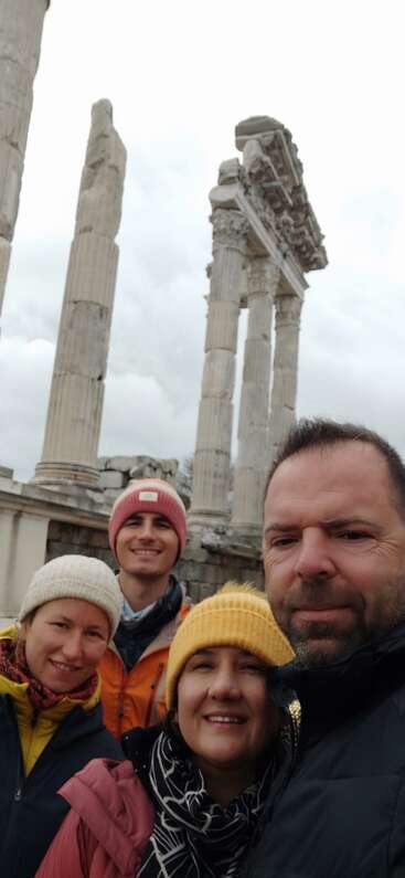 Four people pose for a selfie in front of ancient, weathered stone columns. They wear winter jackets and hats, smiling on a cloudy day at historic ruins.